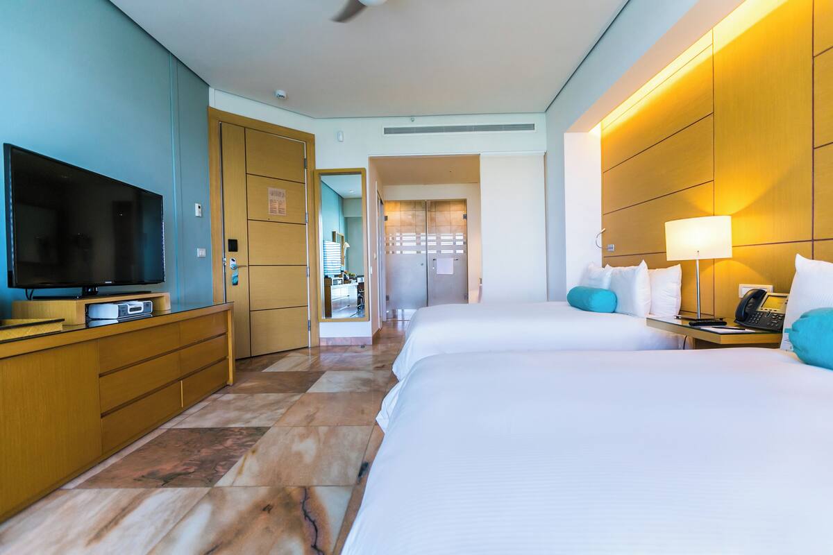A hotel room with a large flat-screen TV, a wooden dresser, two beds with white linens, a bedside table with a lamp, and a telephone.
