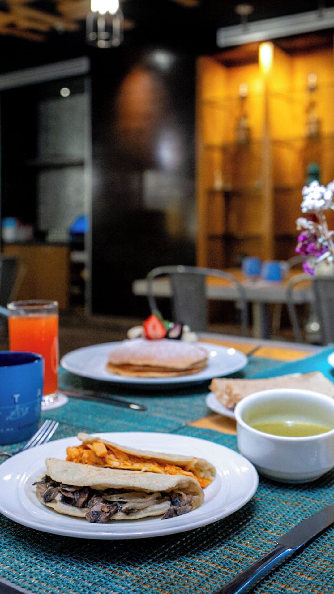 A table with a meal consisting of a folded tortilla with meat, a plate with a folded tortilla and a slice of fruit, and a glass of orange juice. There is a bowl of sauce and a fork on the table.
