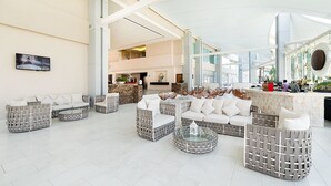 Un hall d’entrée spacieux au design moderne, comprenant un espace salon central avec des canapés blancs et des fauteuils en osier, un comptoir de réception et un grand mur de verre offrant une vue sur l’intérieur.