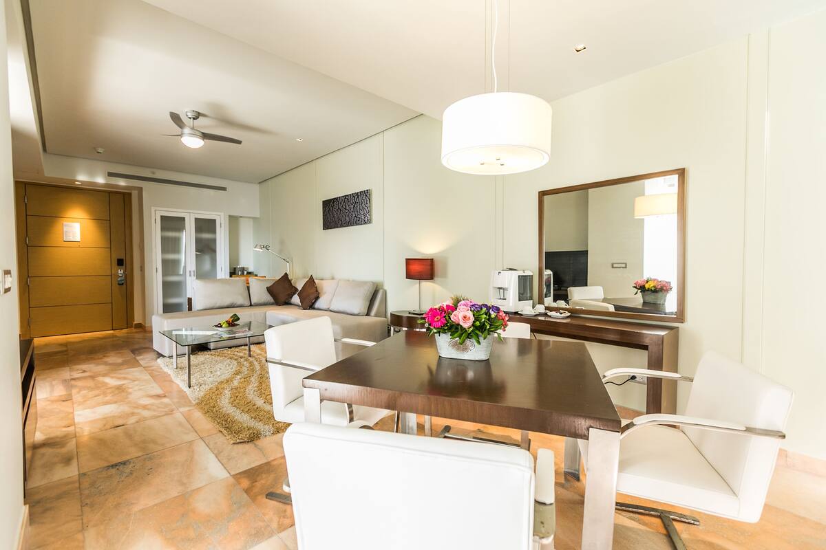 A modern living room with a dining area, a ceiling fan, a large mirror, and a flower vase on the table.