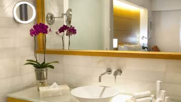 A modern bathroom with a large mirror, white sink, and a wooden vanity. There are purple orchids in a metal pot on the vanity, and a basket of white towels below the mirror.