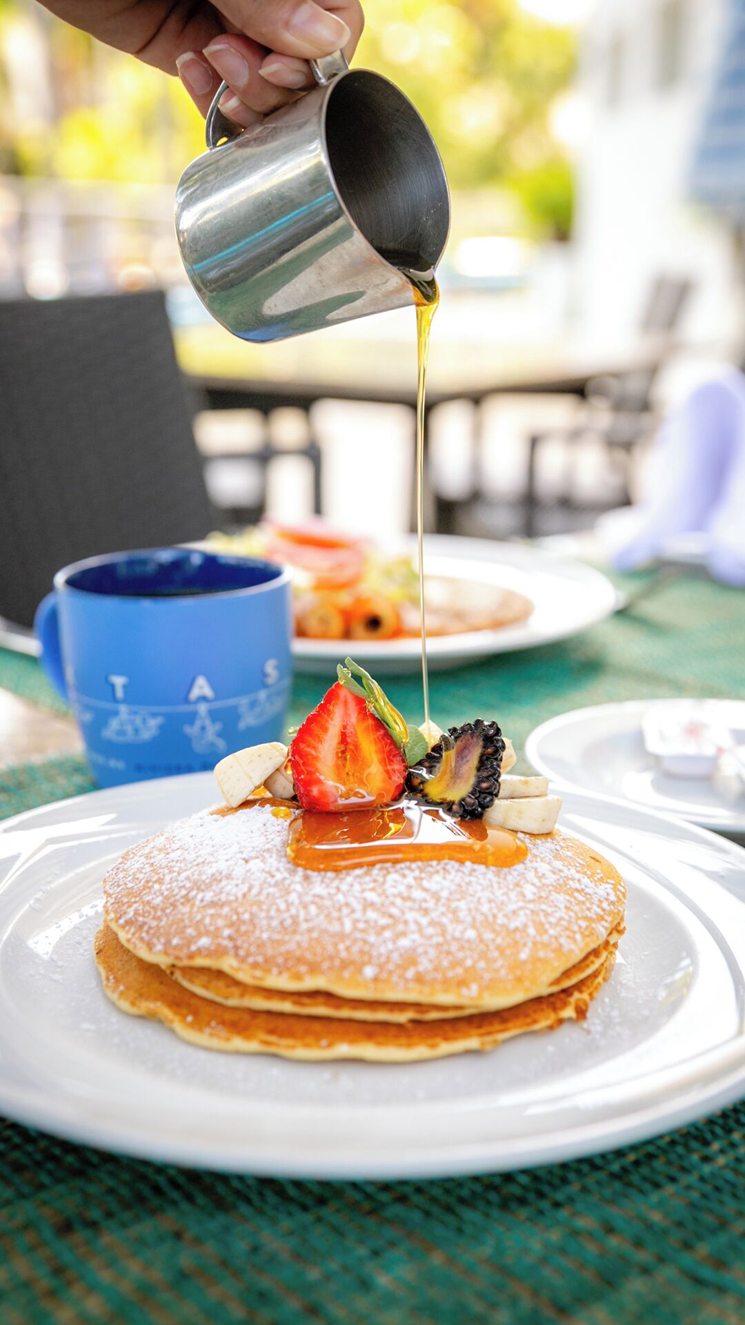 Une pile de crêpes arrosées de sirop, garnies d’une fraise et d’un morceau de fruit.