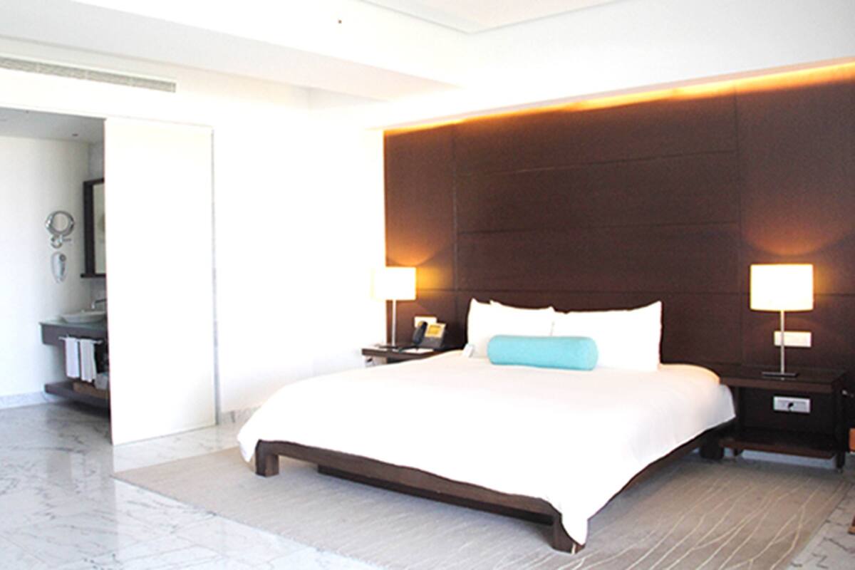 A modern hotel room with a large bed, two bedside tables with lamps, a bathroom visible through an open door, and a marble floor.