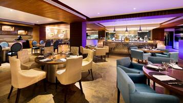 A modern restaurant with round tables and cushioned chairs, a bar area, and a kitchen in the background.