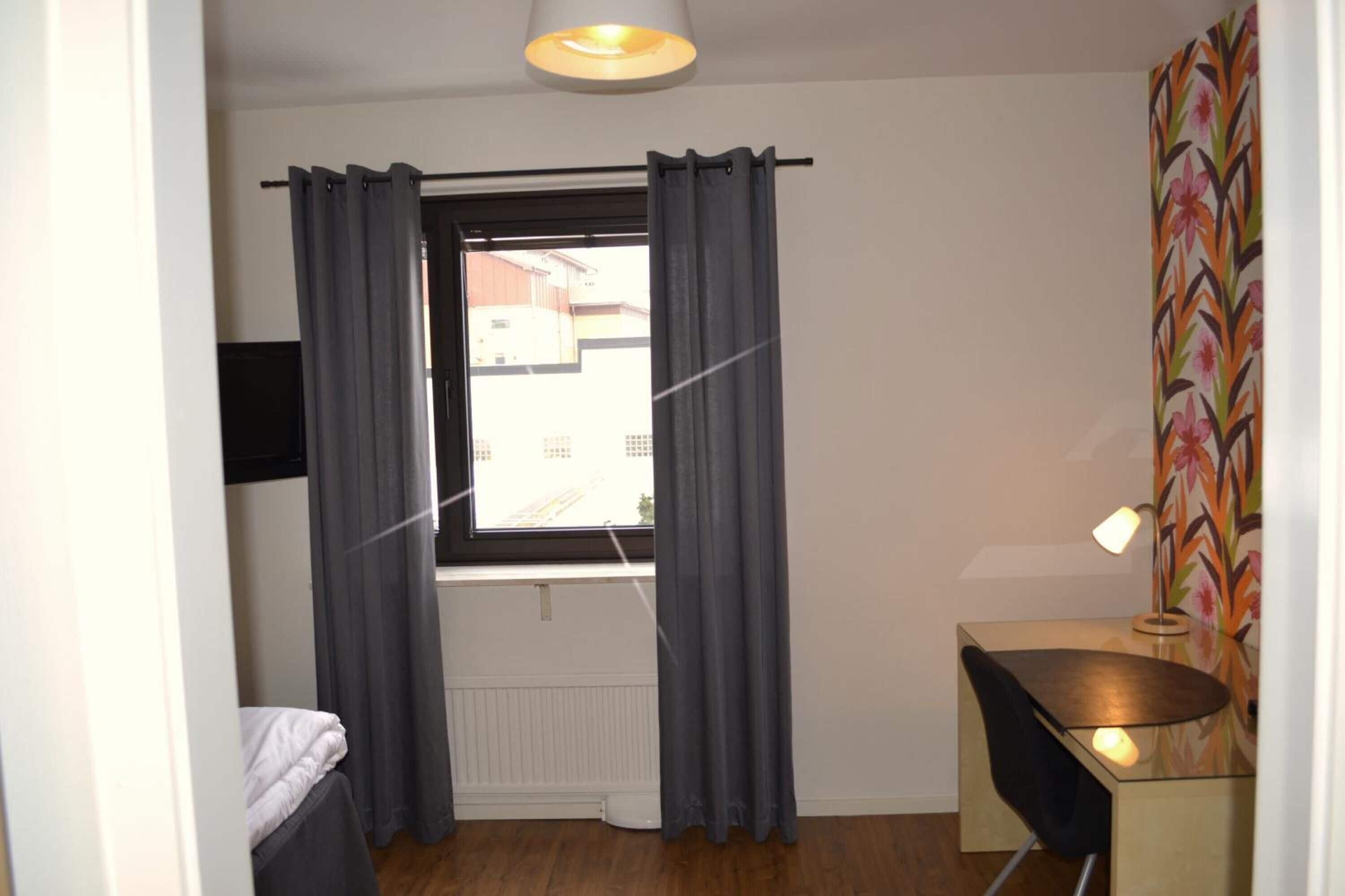 A small room with a bed, a desk, a chair, a window with curtains, and a radiator.