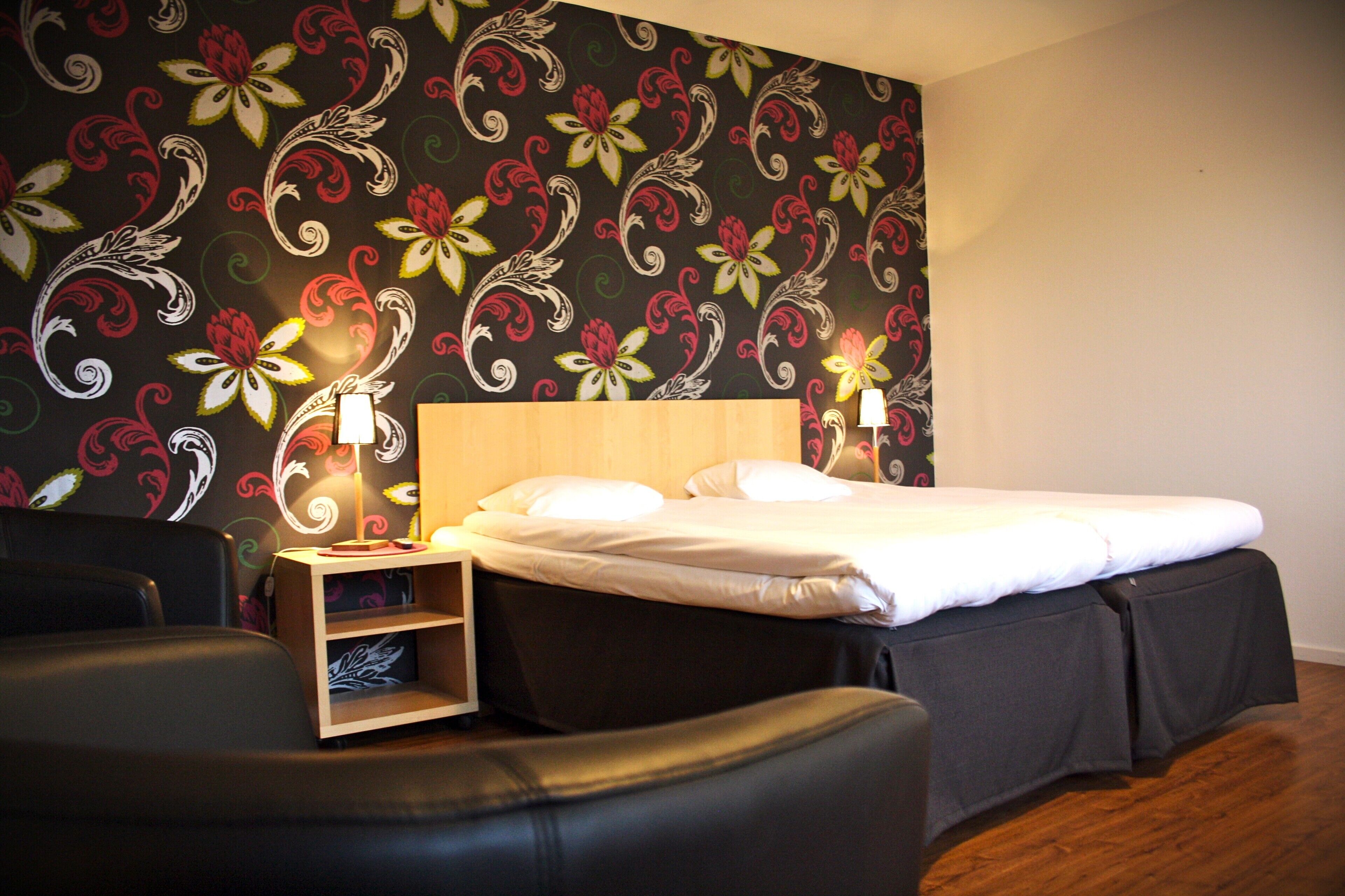 A hotel room with a bed, two bedside tables, a chair, and a decorative floral wallpaper.