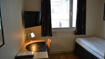 A small, tidy room with a single bed, a desk with a lamp, a TV mounted on the wall, and a window with curtains.