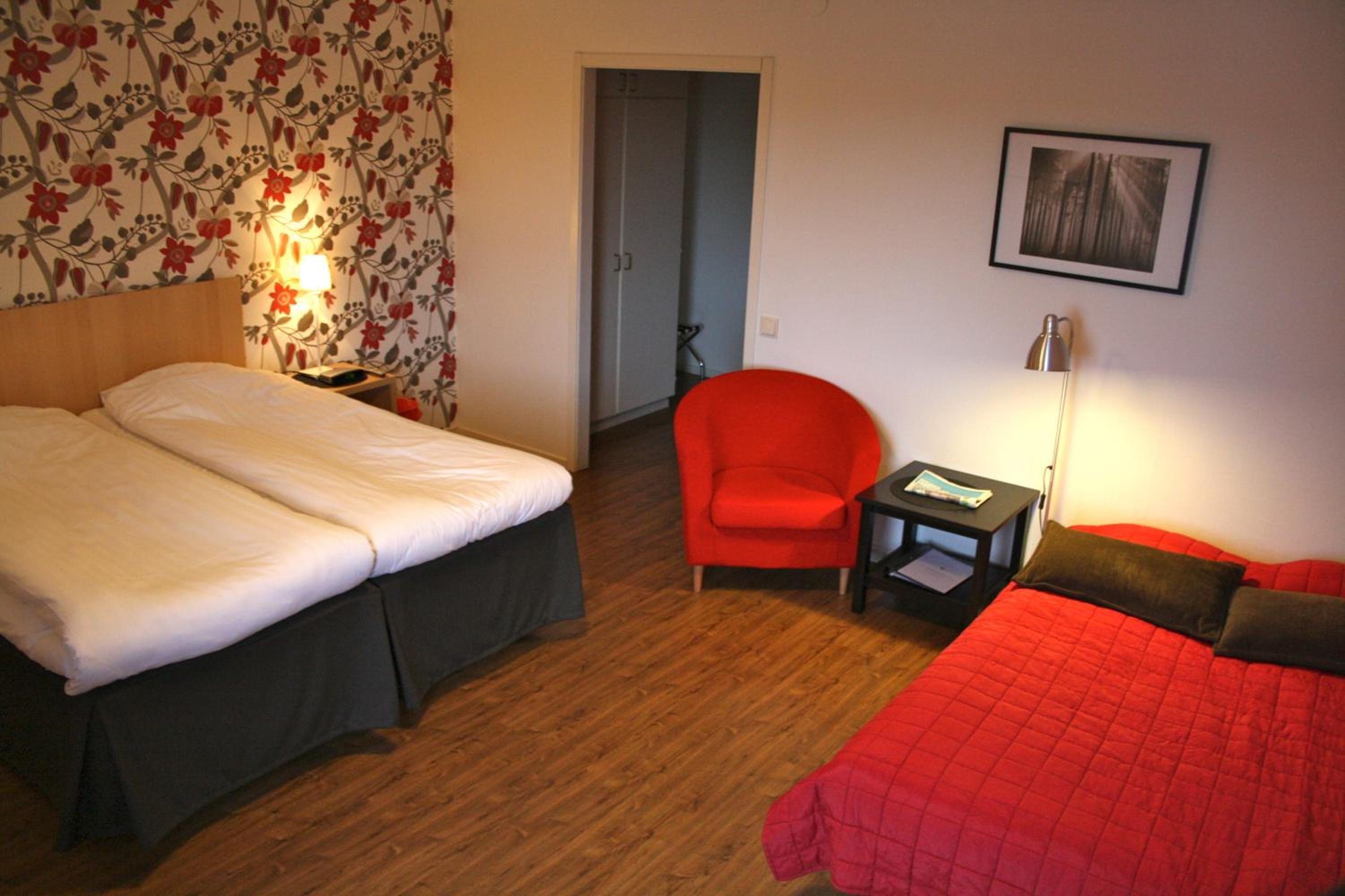A hotel room with two beds, a red chair, a small table, and a framed picture on the wall.