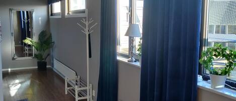 Una habitación con suelo de madera, un perchero blanco, una planta en maceta y una ventana con cortinas azules.