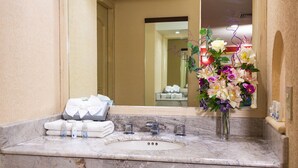 Un baño con lavamanos de mármol, espejo, artículos de aseo y un arreglo floral.