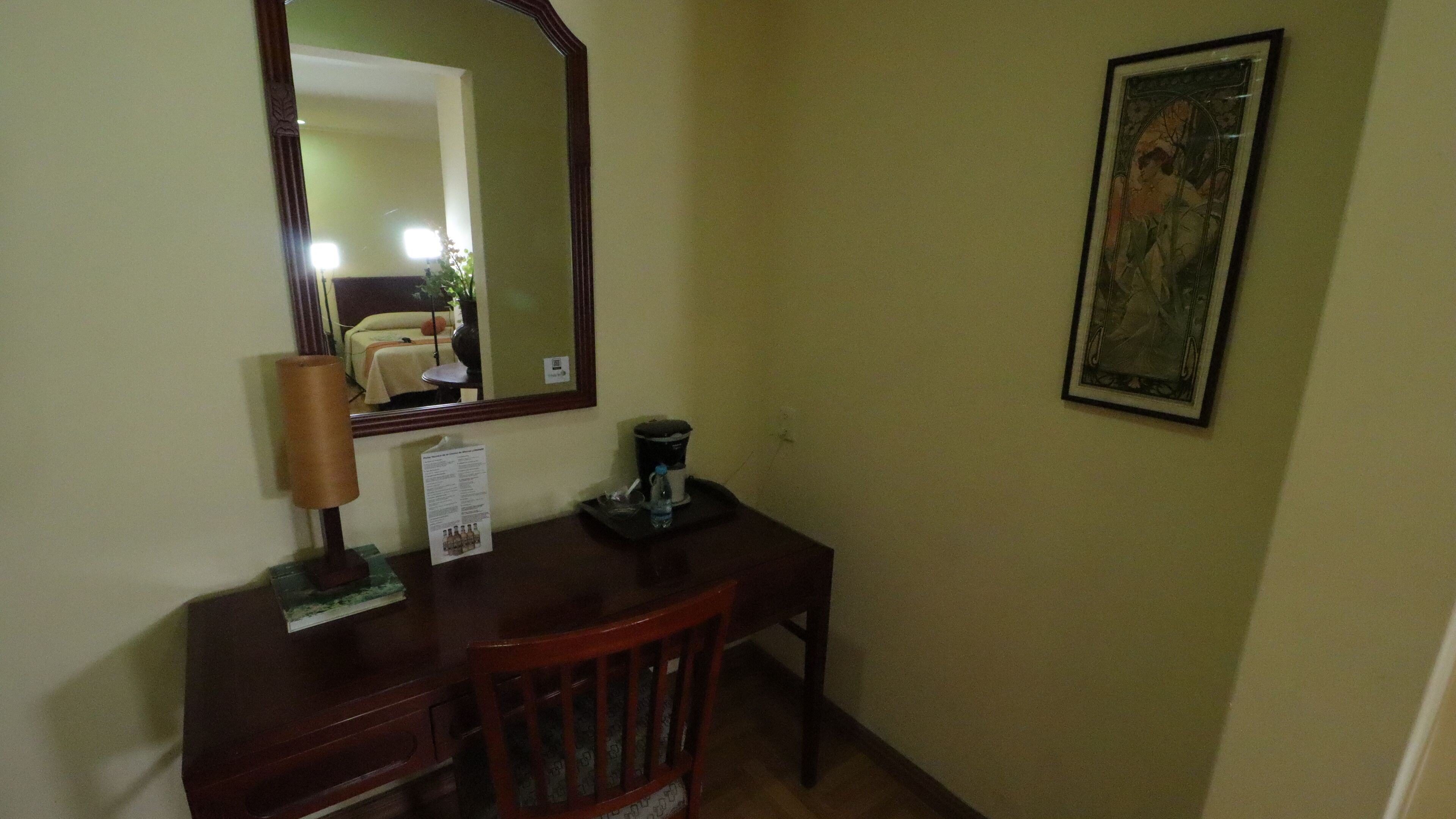 A room with a wooden desk, a mirror, a framed picture on the wall, and a small table with a water bottle and a coffee maker.