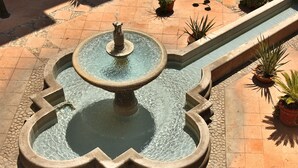 Uma fonte em formato de coração com um bocal central, rodeada por um pátio com azulejos e plantas em vasos.