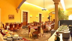 Uma área de refeições com várias mesas e cadeiras, um buffet com diversos pratos e uma planta grande ao fundo.