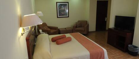 A hotel room with a bed, two chairs, a television, and a painting on the wall.
