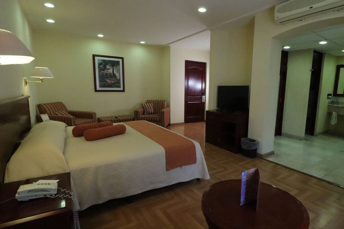 Habitación de hotel con una cama, dos sillas, una mesita, un televisor y un baño visible a través de una puerta abierta.