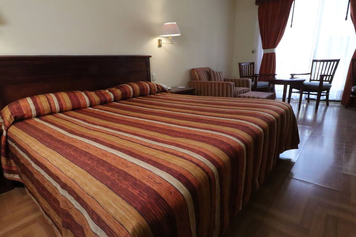 Una habitación de hotel con una cama grande, dos mesitas de noche, un escritorio con una silla y una ventana con cortinas.