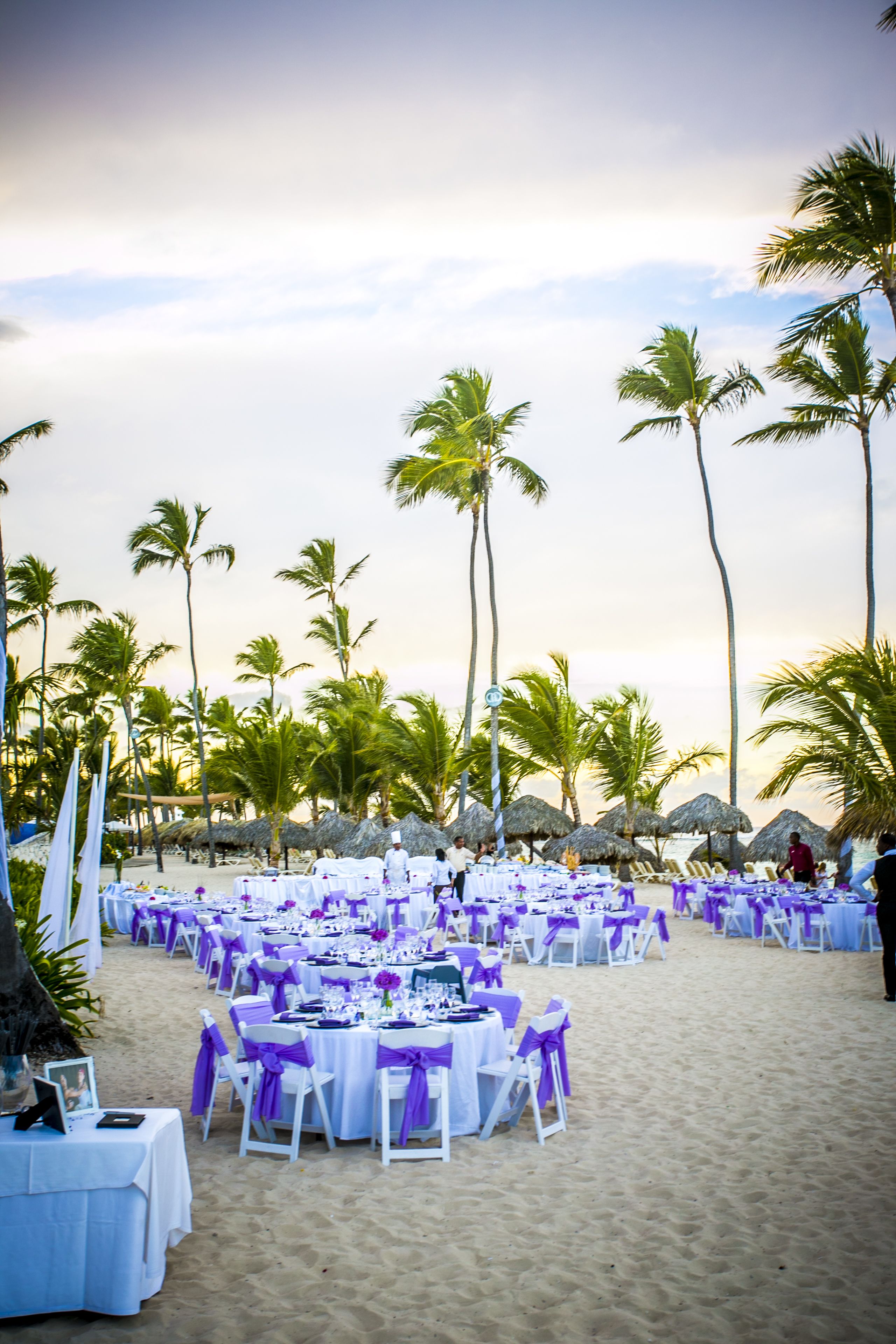 A beachside event setup with tables and chairs arranged on the sand, surrounded by palm trees.