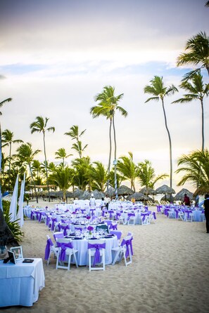 A beachside event setup with tables and chairs arranged on the sand, surrounded by palm trees.