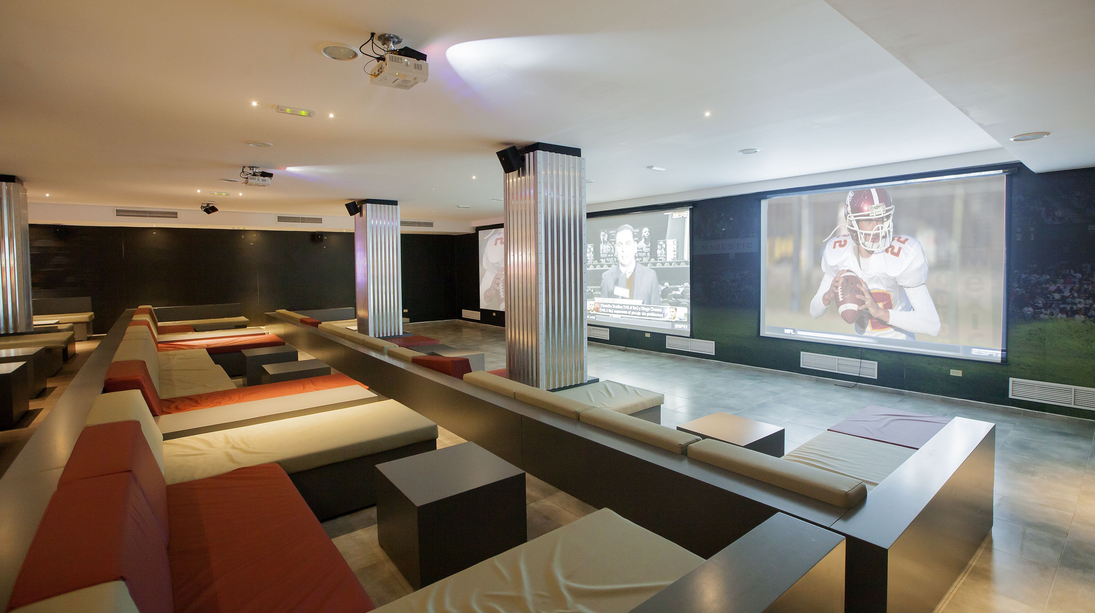 A modern hotel lobby with tiered seating, a large screen displaying sports content, and a ceiling with recessed lighting.