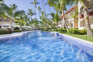 A clear blue pool surrounded by palm trees and a resort building with multiple balconies. - Majestic Elegance Punta Cana - All Inclusive (Punta Cana)