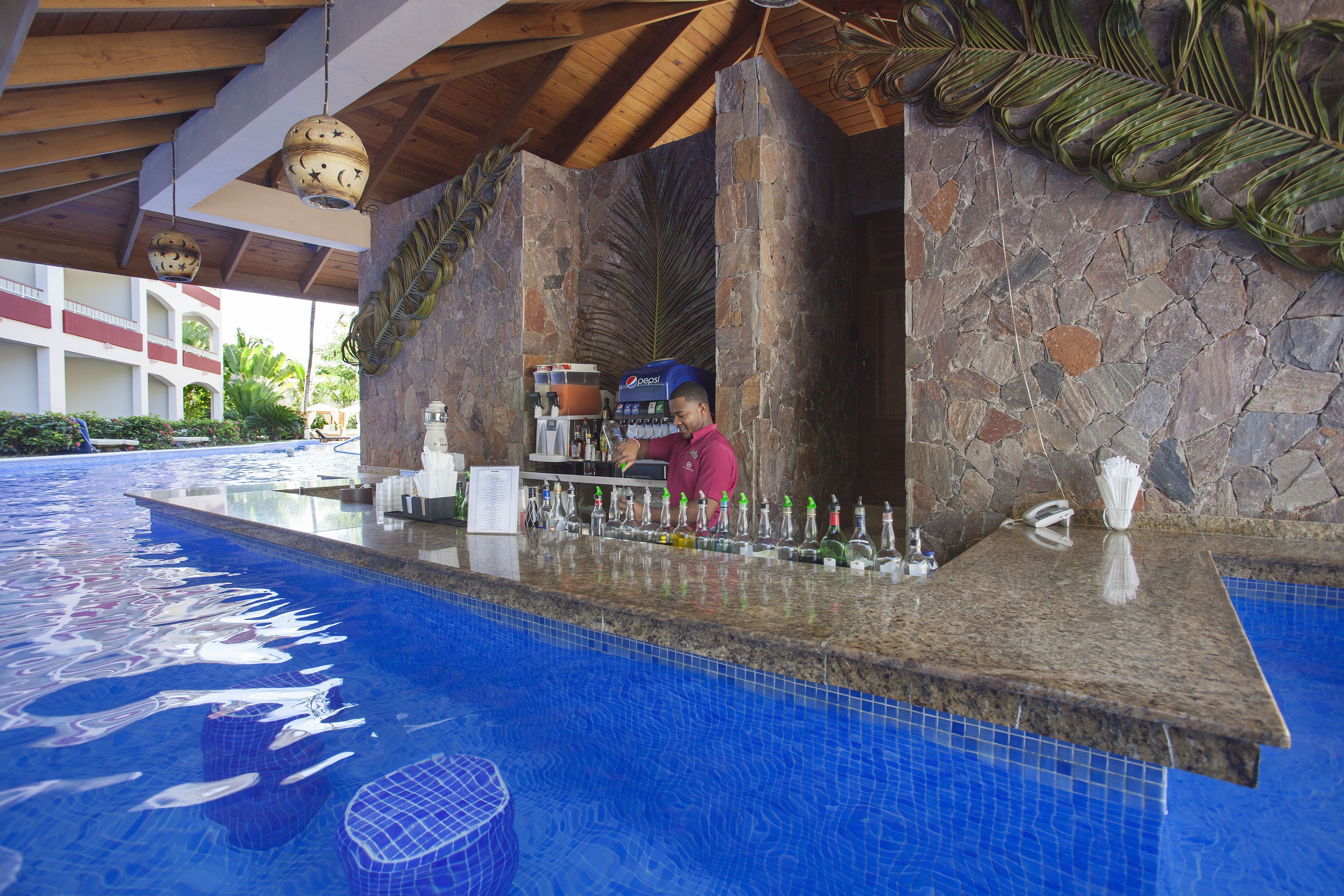 Un bar junto a la piscina con un fondo de pared de piedra, una barra y una persona sirviendo bebidas.