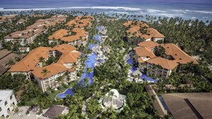 Vista aérea de un complejo turístico con varias construcciones, una piscina central y palmeras.