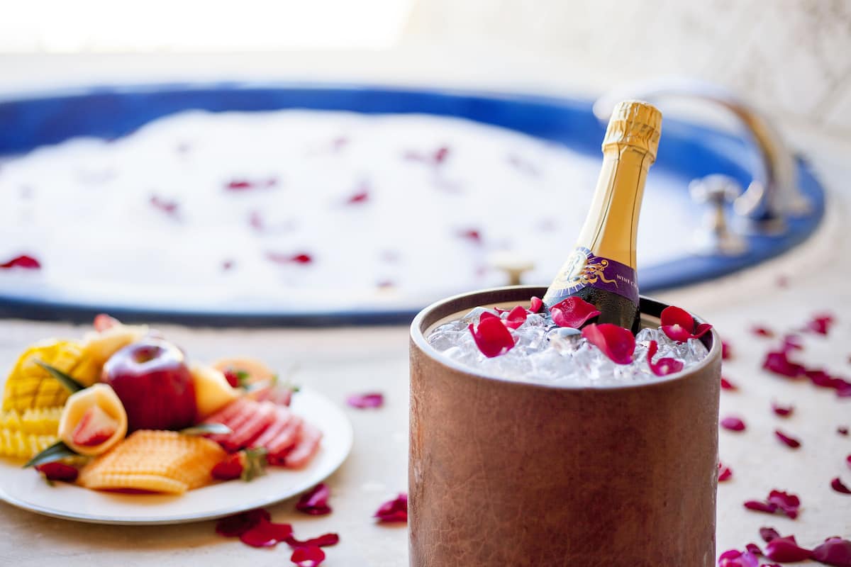 Eine Flasche Champagner in einem Kupferbecher mit Eis und Rosenblüten, ein Obstteller und eine Badewanne im Hintergrund.