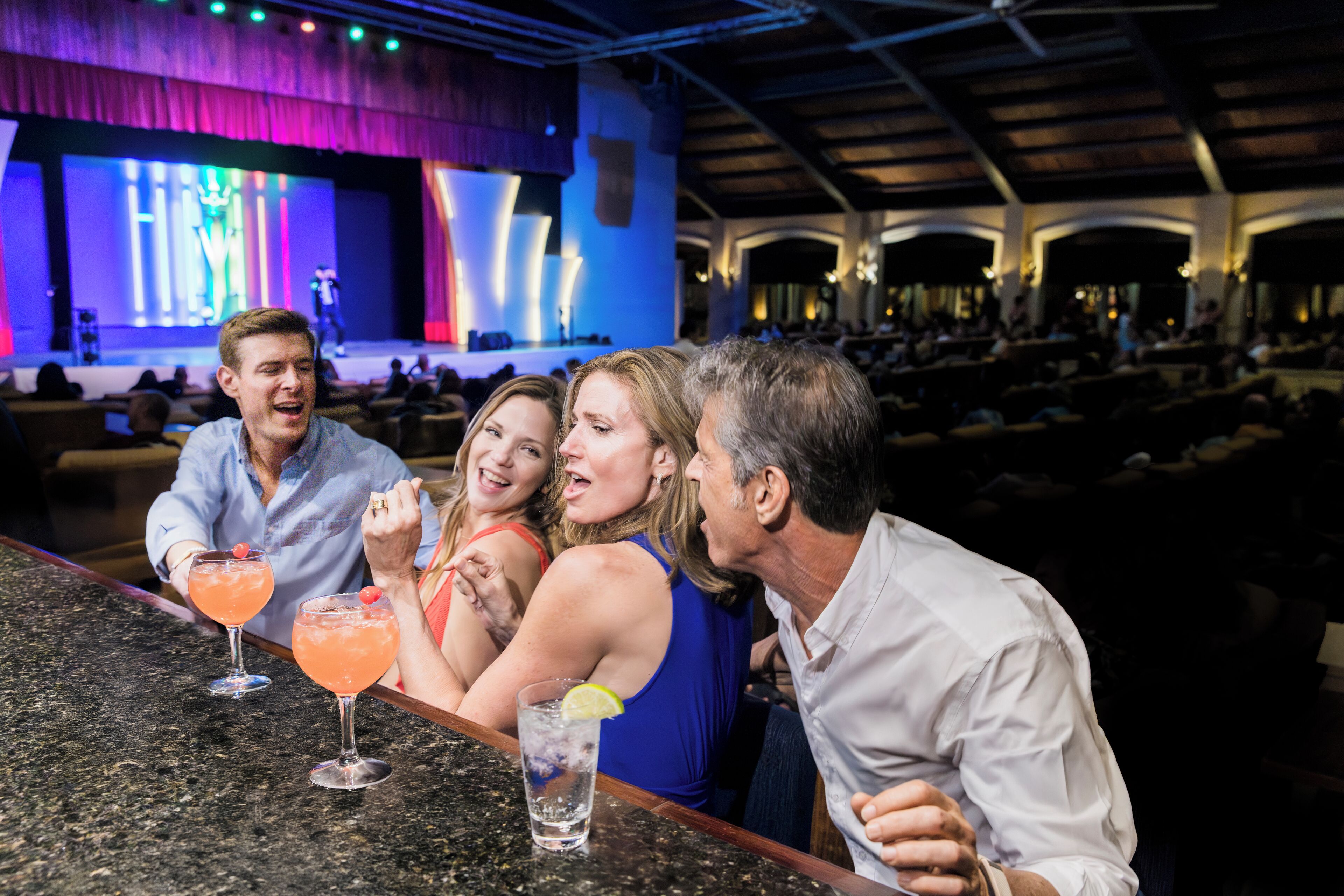 A bar area with a stage in the background, seating for guests, and drinks on the counter.
