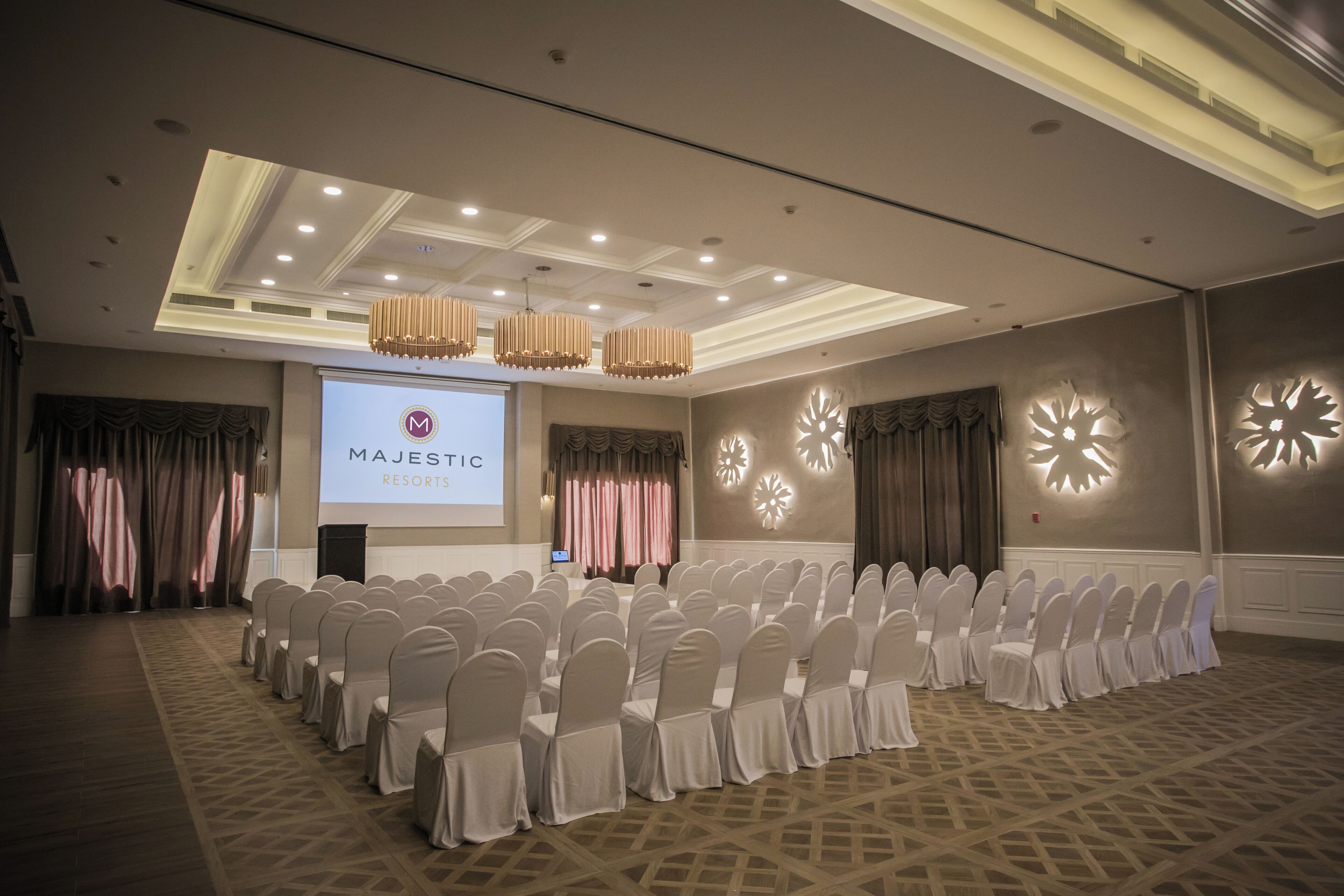 Een congreszaal bij Majestic Resorts met rijen witte stoelen, een groot scherm waarop het logo van het resort staat en decoratieve wandlampen.