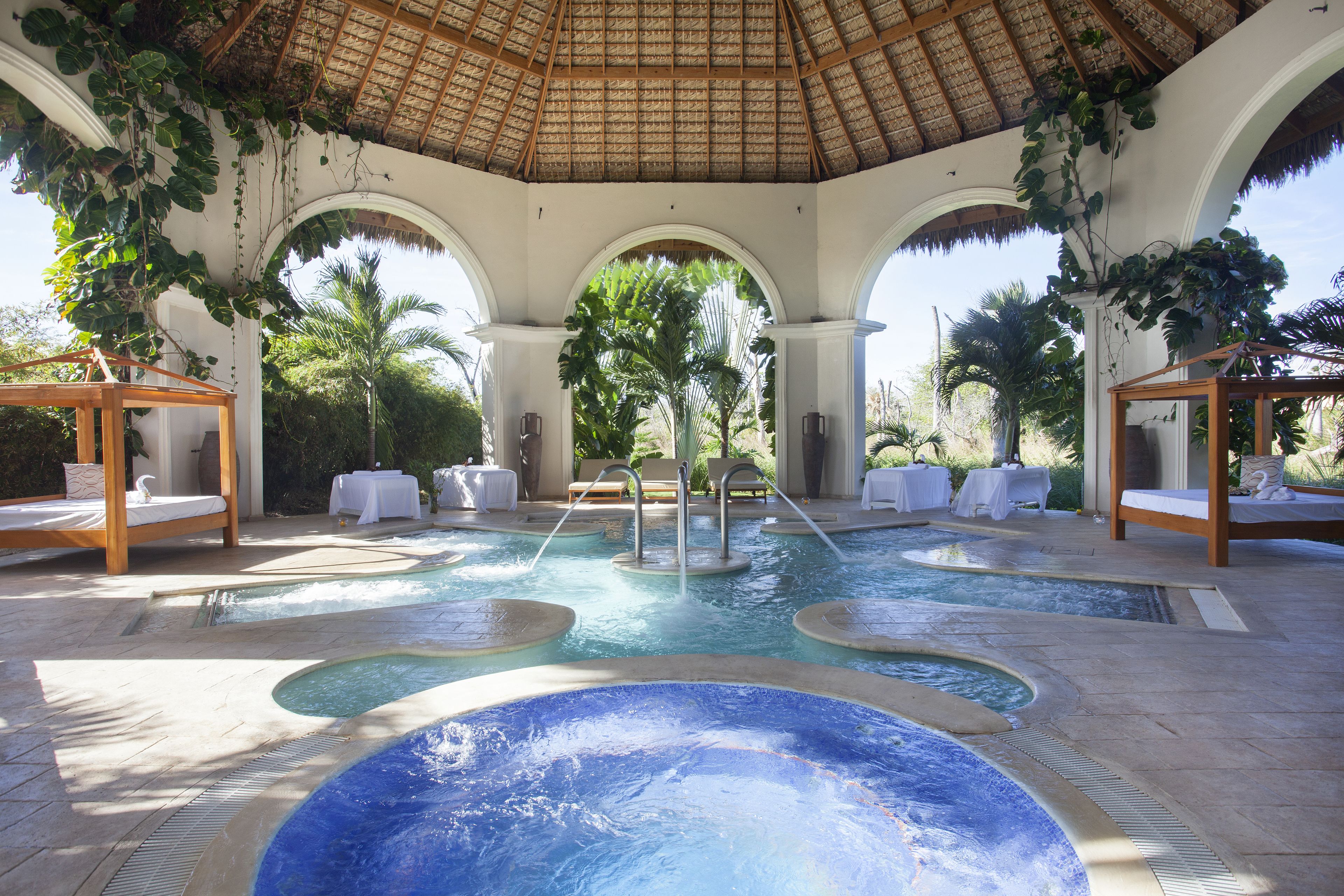 A pool area with multiple hot tubs, lounge chairs, and a thatched roof structure.