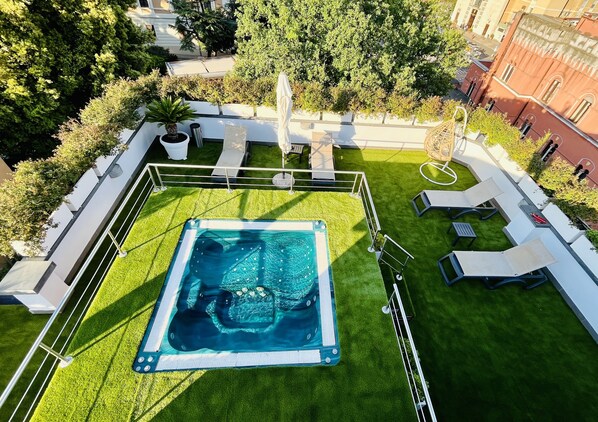 Vue aérienne d’un espace piscine sur le toit, avec un jacuzzi, entouré de gazon synthétique vert, de rampes blanches et de chaises longues.