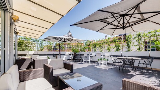 Un patio al aire libre con mobiliario de mimbre, sombrillas grandes y vistas de edificios al fondo.
