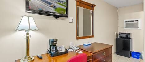 A hotel room with a desk, a television mounted on the wall, a mirror, a coffee maker, a telephone, a microwave, and a refrigerator.