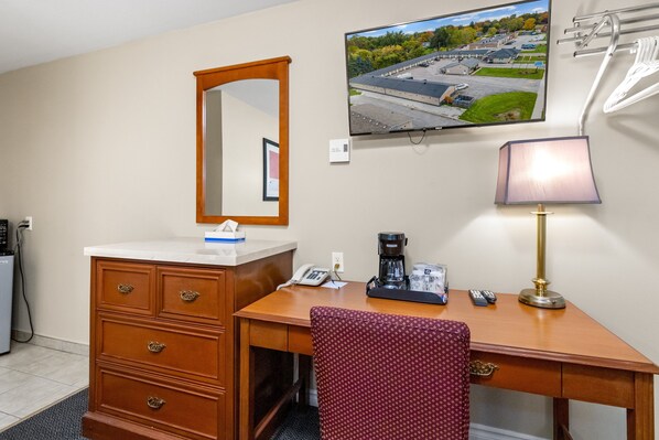 A hotel room with a wooden desk, a chair, a mirror, a television, a lamp, a coffee maker, and a telephone.