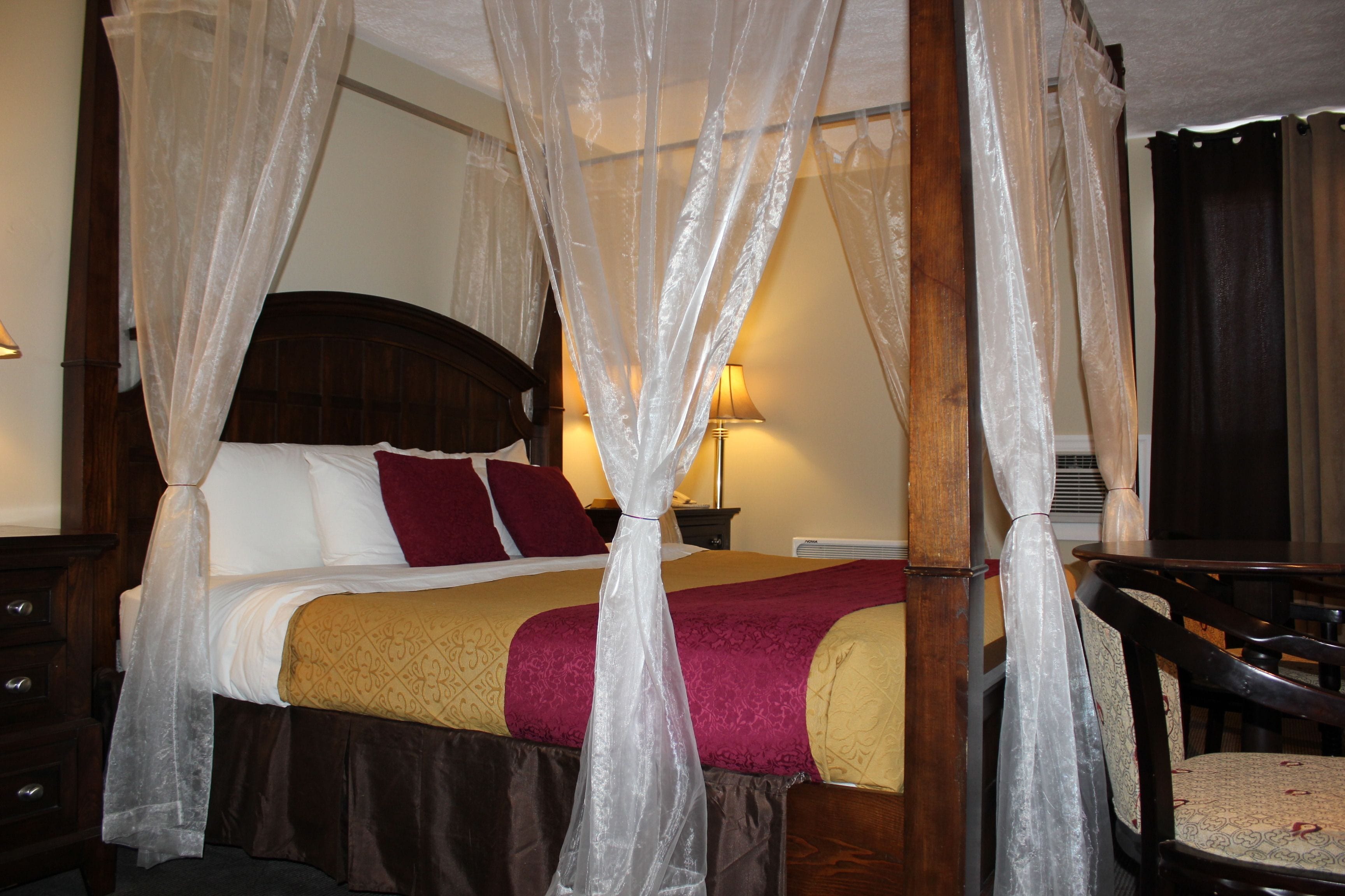 A four-poster canopy bed with a wooden headboard, a red and gold bedspread, and a white bed skirt.