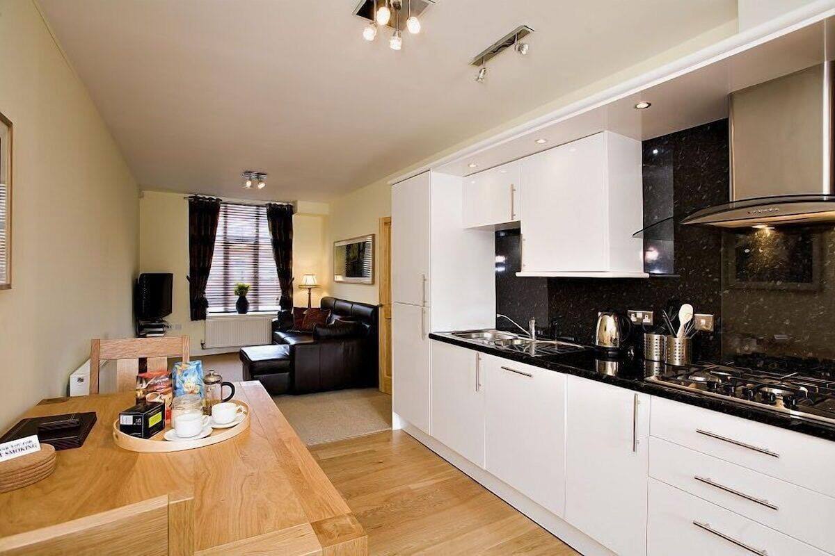 A modern kitchen with white cabinets, a black countertop, and stainless steel appliances. There is a dining area with a wooden table and chairs, and a living room with a sofa and a TV.