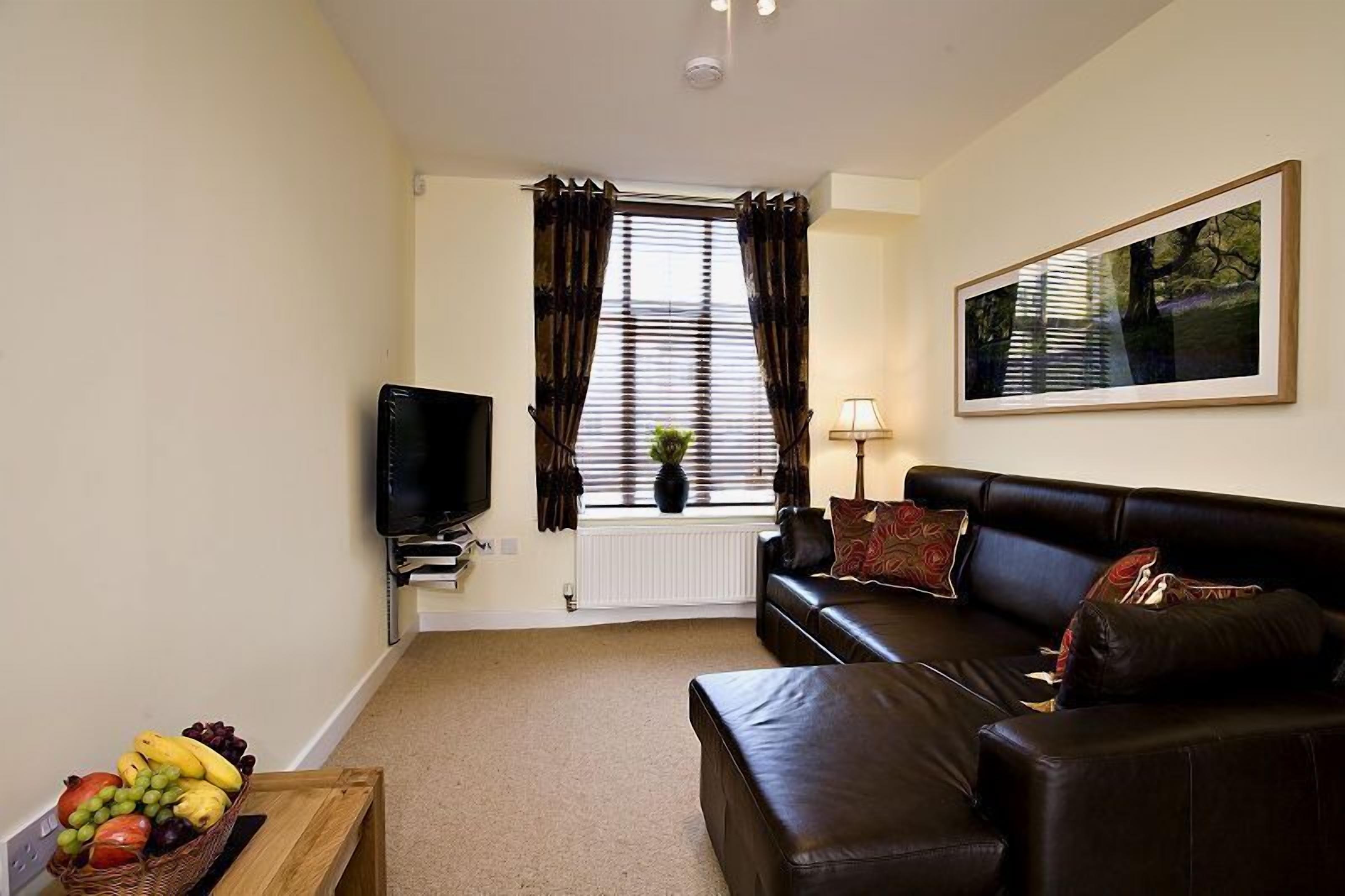 A living room with a black leather sofa, a television, a wooden coffee table with fruit, and a window with curtains.