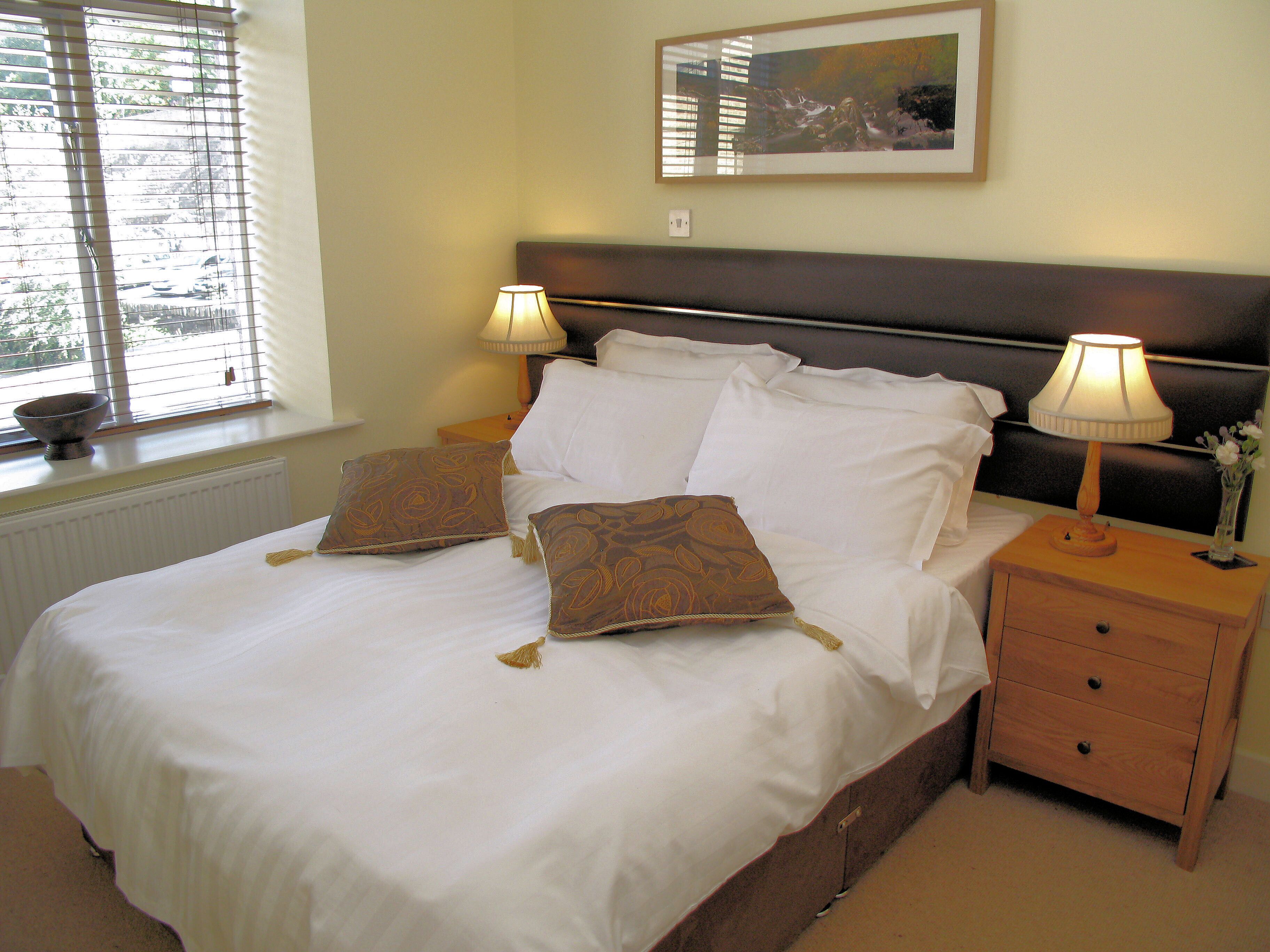 A neatly made bed with white linens and two decorative pillows, a bedside table with a lamp, and a framed artwork on the wall.