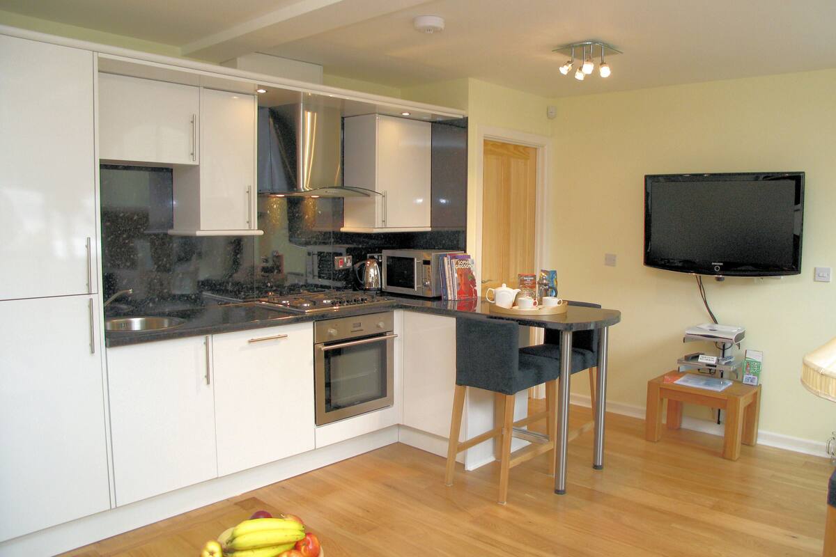 A modern kitchen with white cabinets, a built-in oven, and a microwave. There is a fruit bowl on the counter and a small table with a TV and a DVD player.