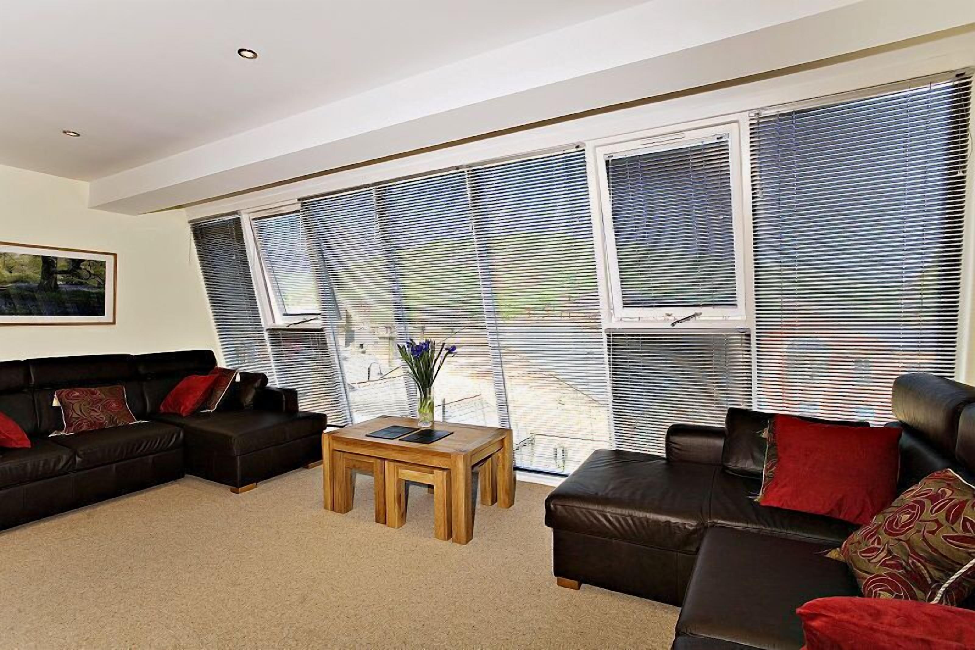 A modern living room with two black leather sofas, a wooden coffee table, and large windows with blinds.