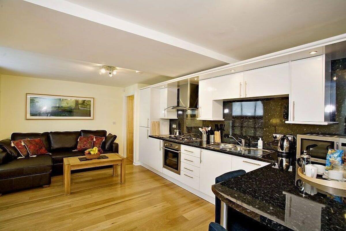 A modern kitchen with white cabinets, a black countertop, and a dining area with a wooden table and a black leather couch.