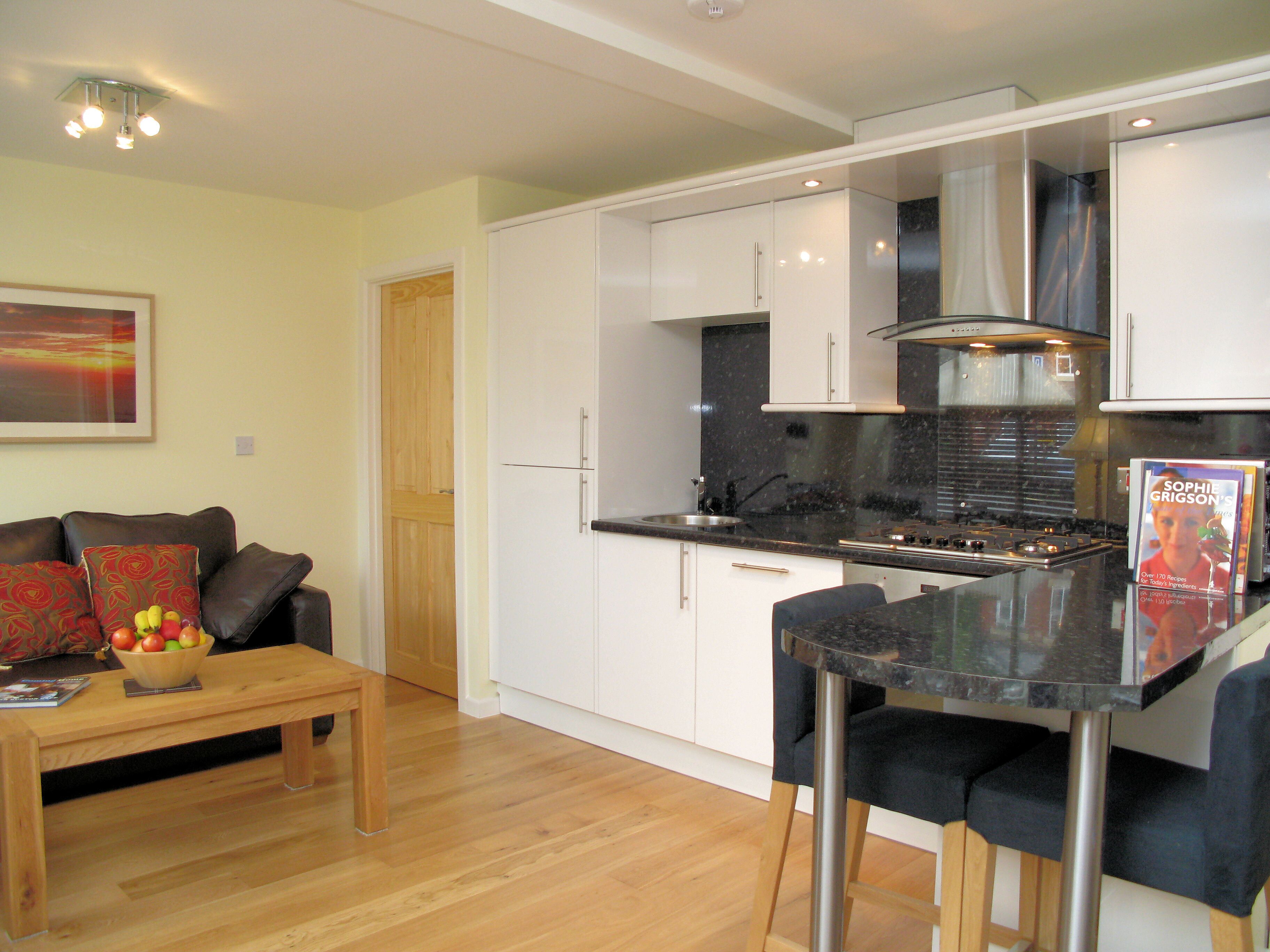 A modern kitchen with white cabinets, a black countertop, and stainless steel appliances. There is a dining area with a wooden table and chairs, and a bowl of fruit on the table.