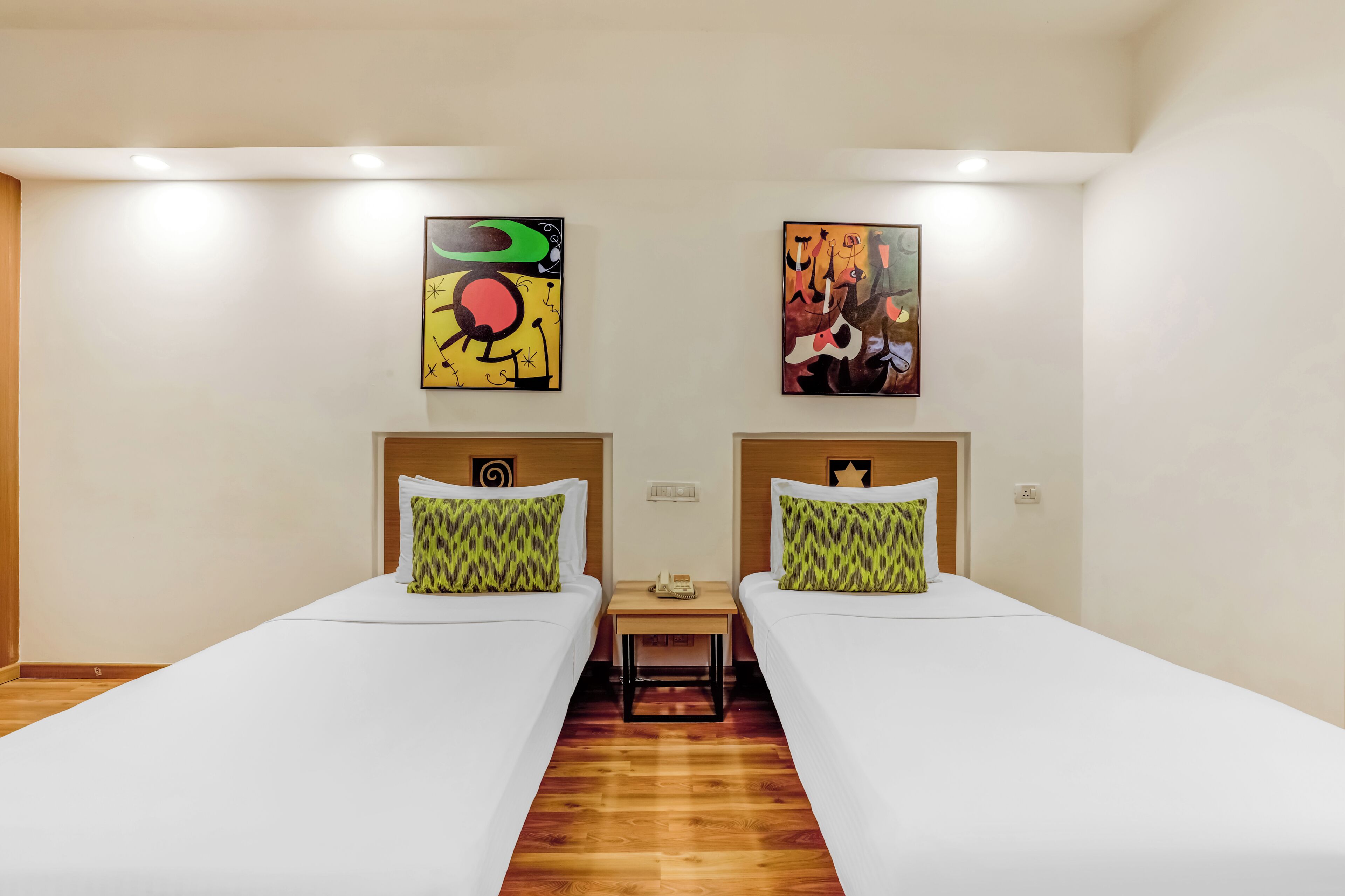 A hotel room with two single beds, a wooden floor, a small table, and two abstract paintings on the walls.