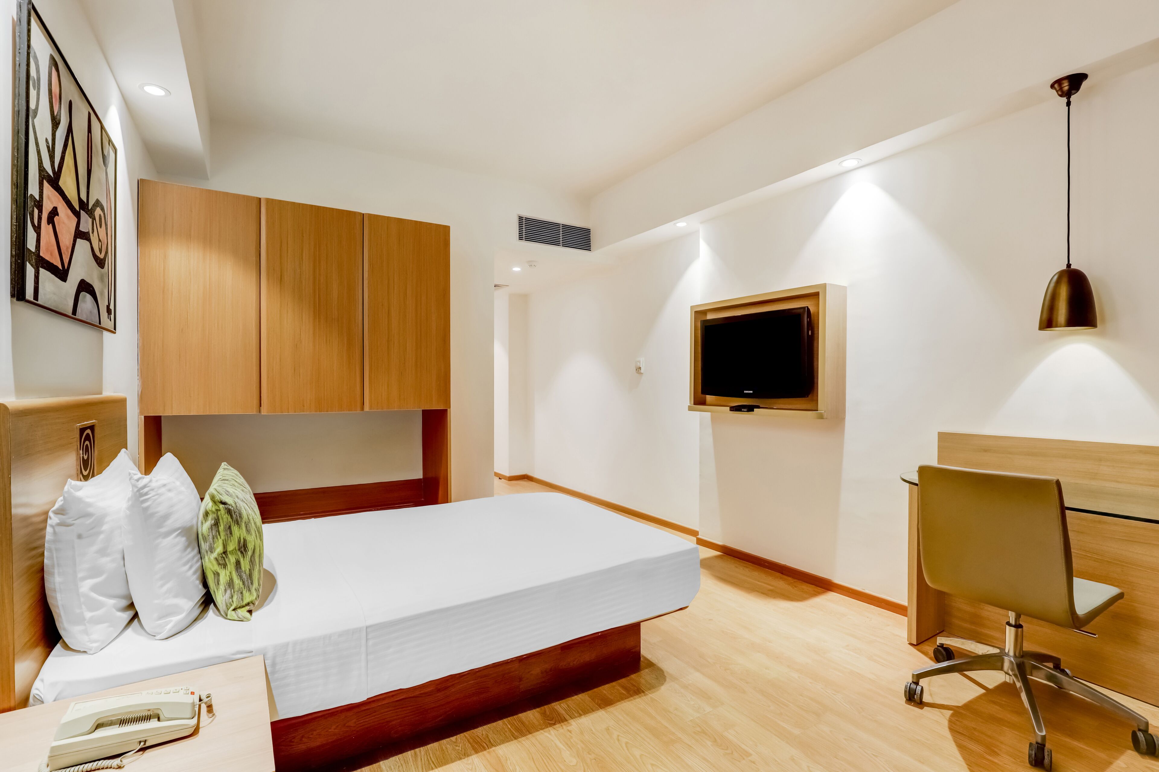 A modern hotel room with a bed, a desk, a chair, a TV, and a painting on the wall.