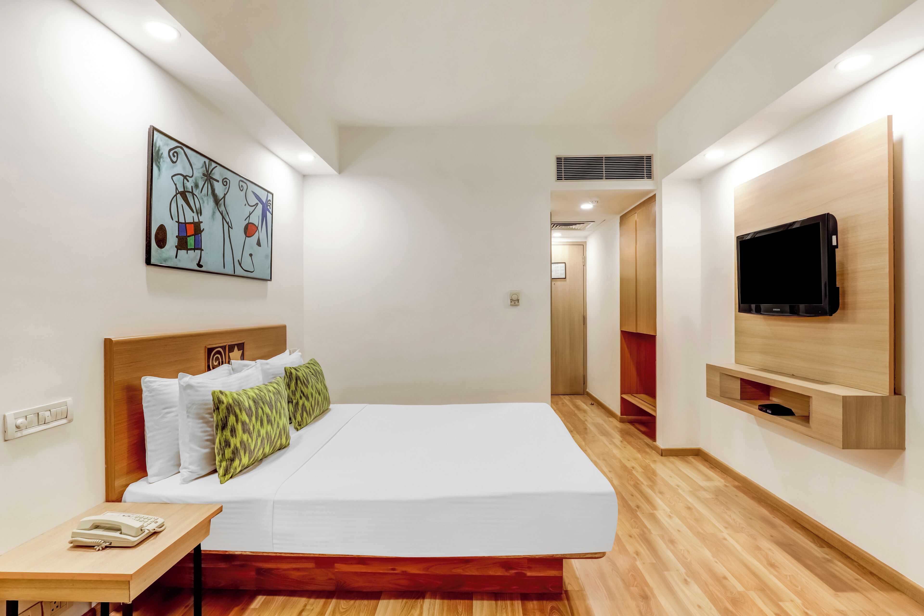A modern hotel room with a wooden bed, a mounted TV, and a painting on the wall.