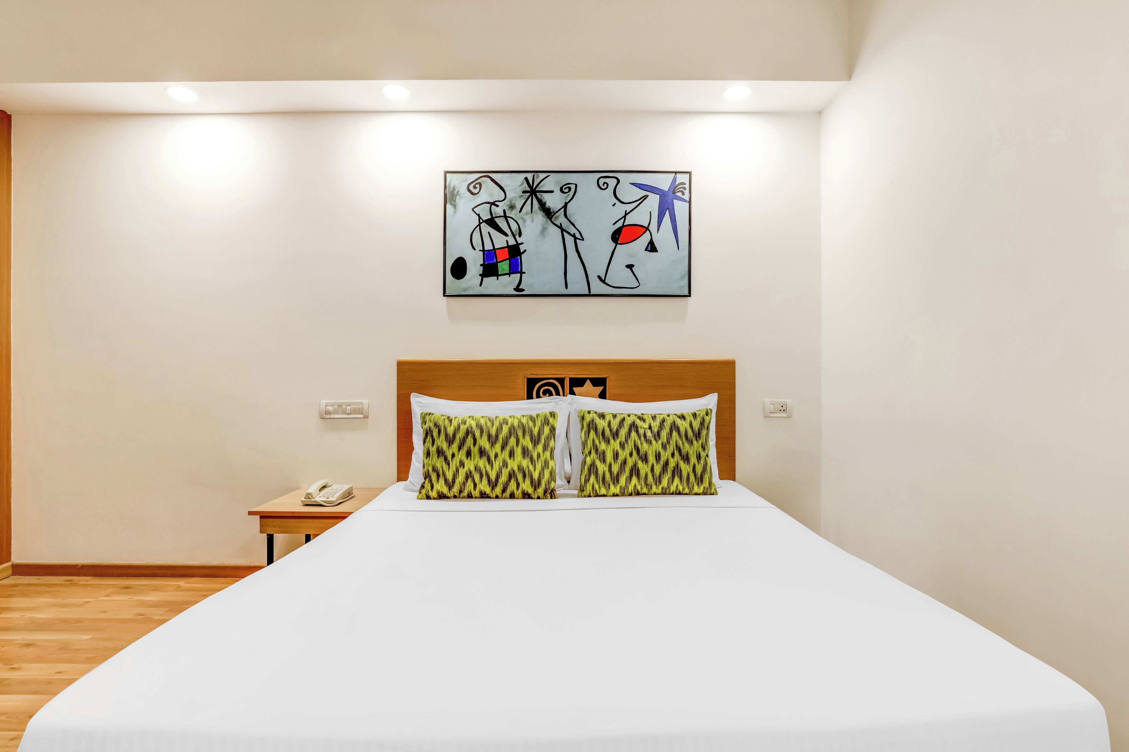A hotel room with a bed, two patterned pillows, a bedside table with a phone, and a wall art piece.