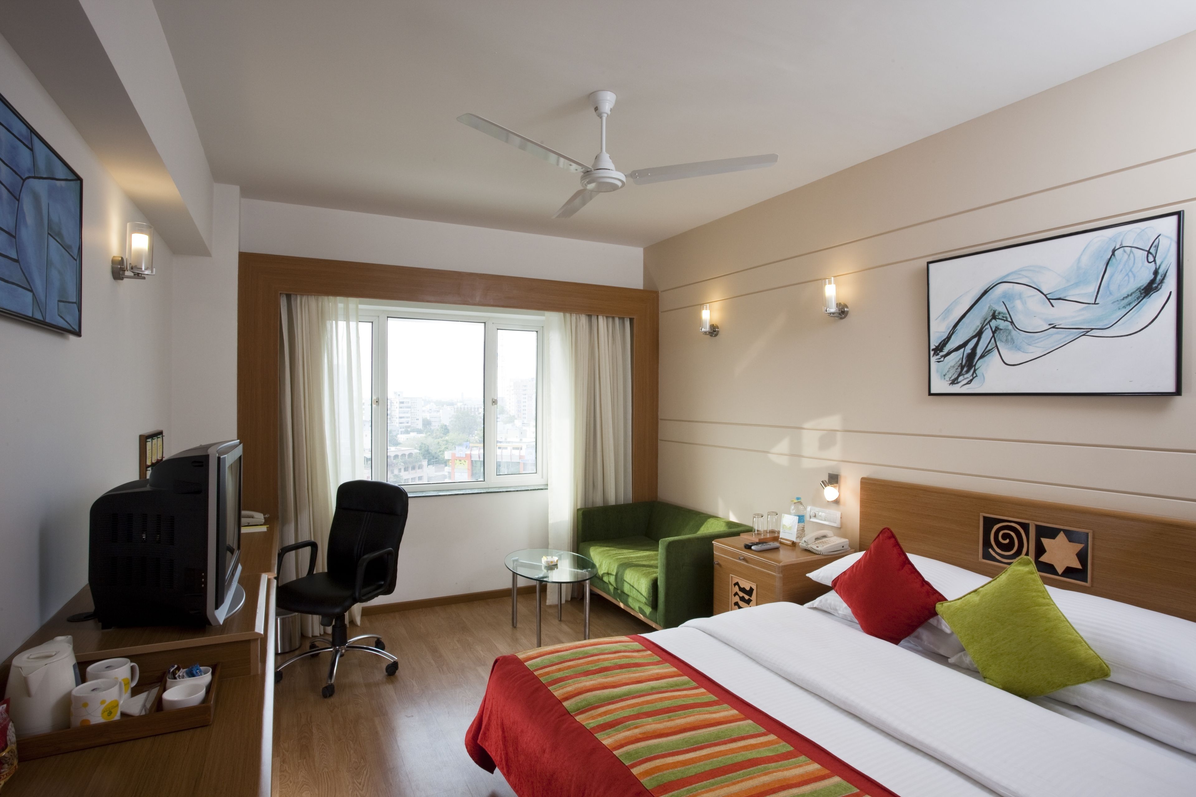 A hotel room with a bed, a desk with a computer, a green armchair, a small table, a window with curtains, and a ceiling fan.
