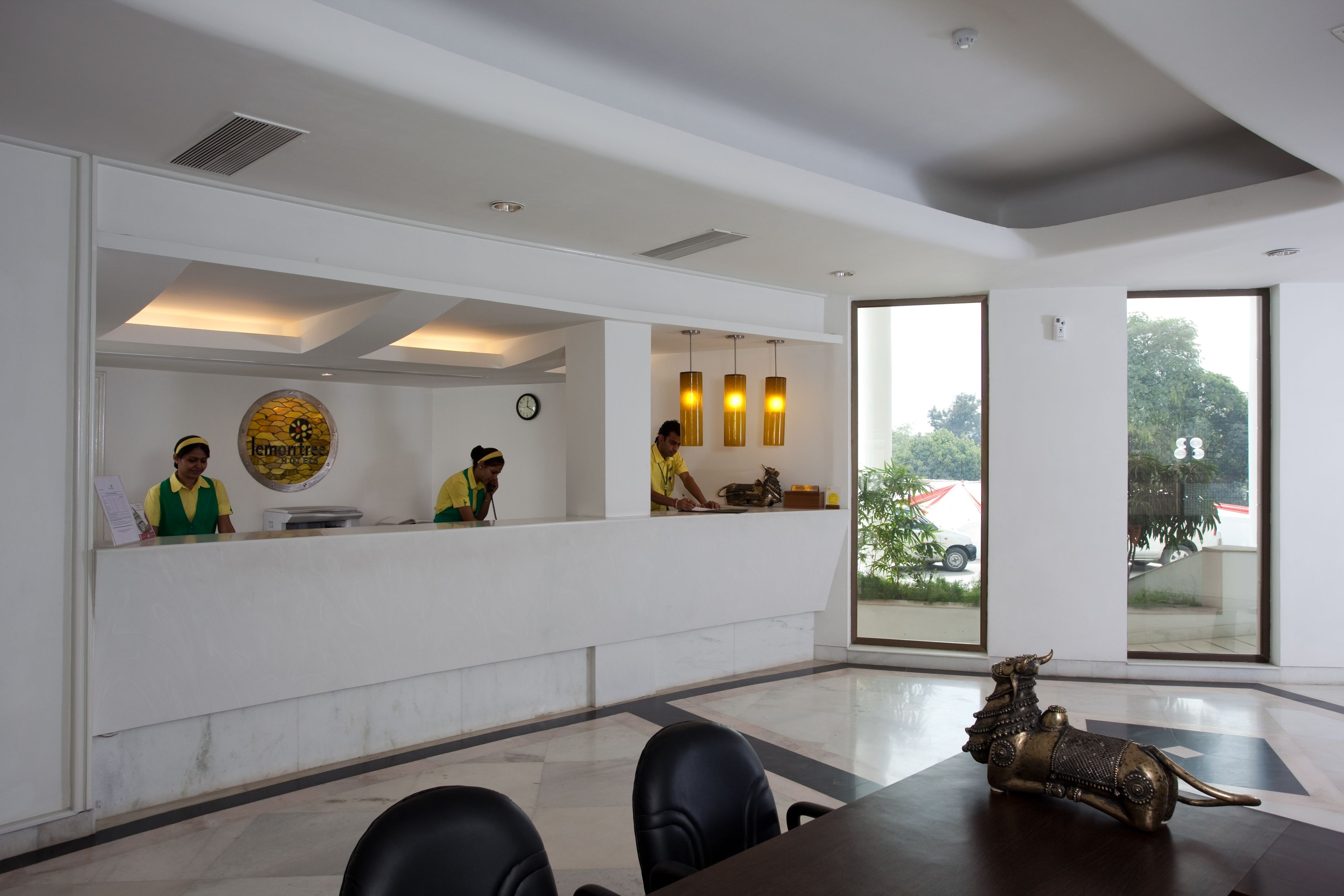 Zona de recepción de un hotel con un mostrador, tres empleados, un ventanal grande y una escultura decorativa sobre la mesa.