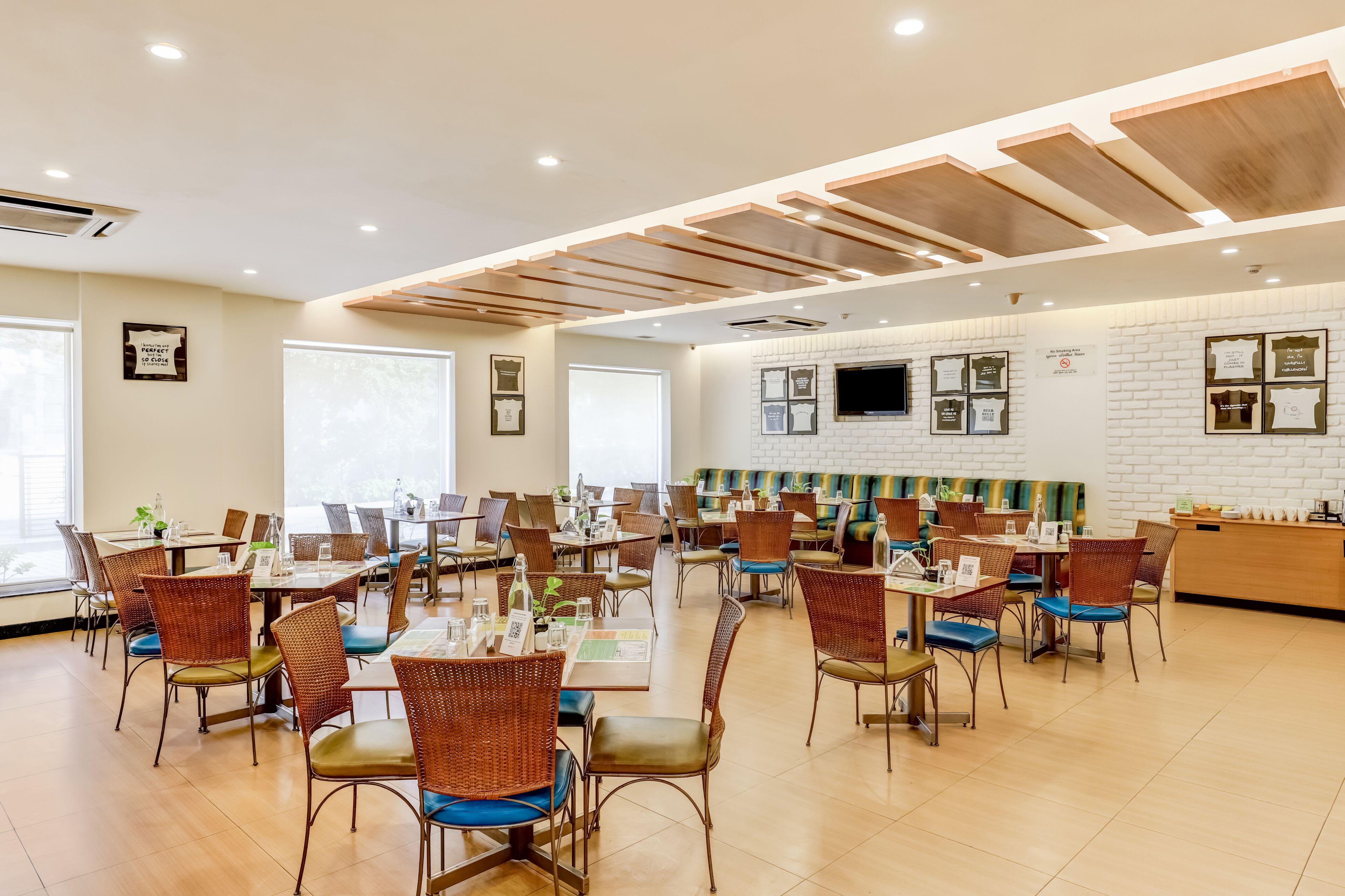 Un vaste espace dédié à la salle à manger, avec de nombreuses tables et chaises, des panneaux de plafond en bois et un mur en briques orné de tableaux encadrés.