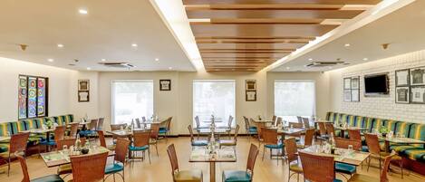 Une salle à manger spacieuse avec de hauts plafonds, des poutres en bois et de grandes fenêtres. Les tables et les chaises sont disposées avec soin, et des coussins colorés sont posés sur les bancs.